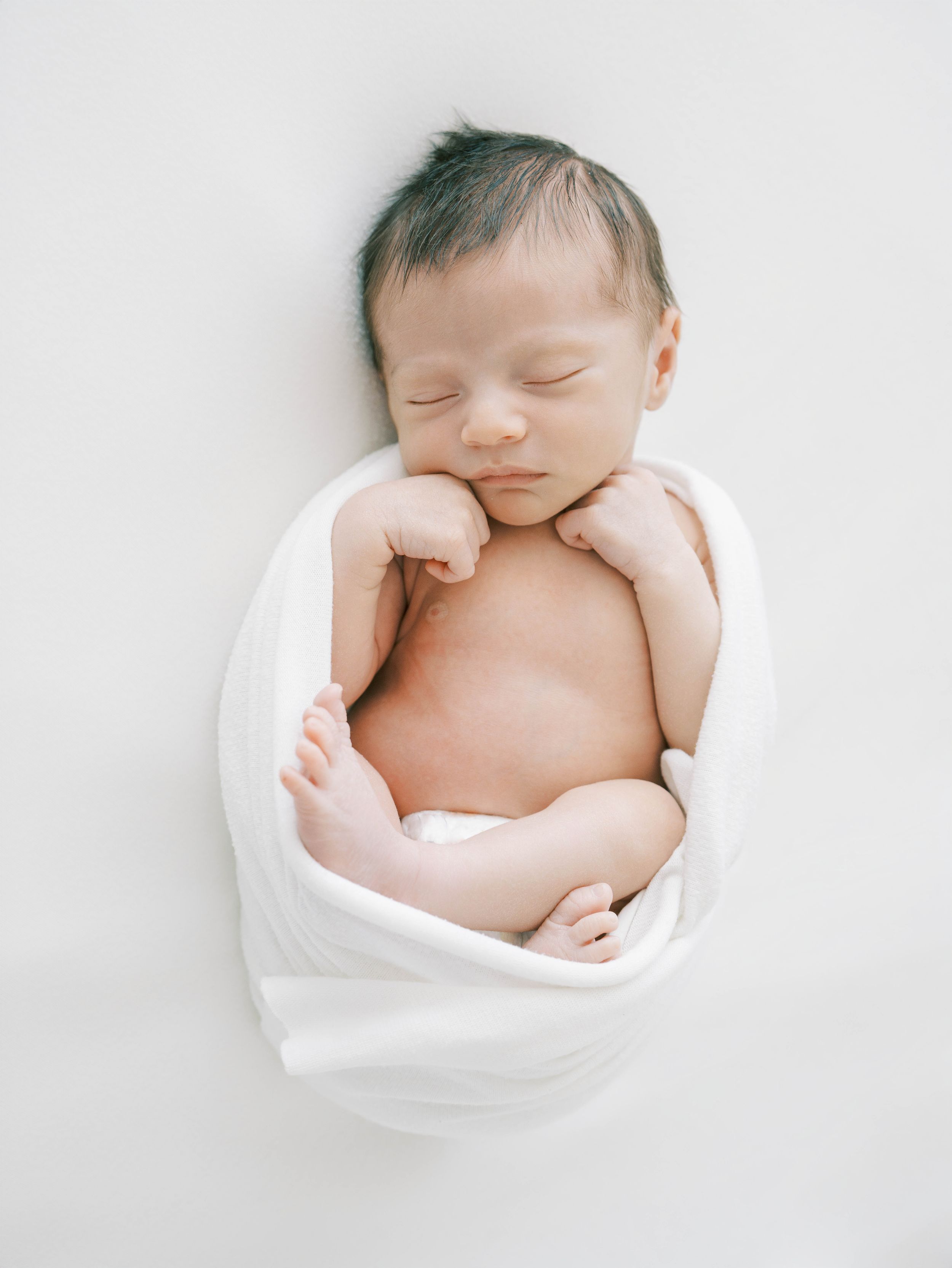 Peaceful sleeping newborn baby wrapped in soft white fabric against light background.