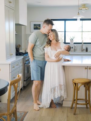 A couple stands together in a bright white kitchen with hardwood floors and rattan bar stools.
