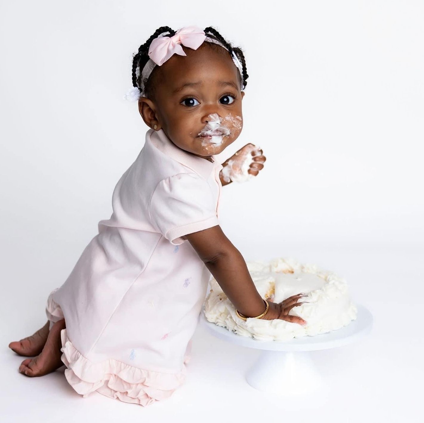 Baby in pink dress plays with white cake frosting while sitting on white backdrop during cake smash photoshoot.