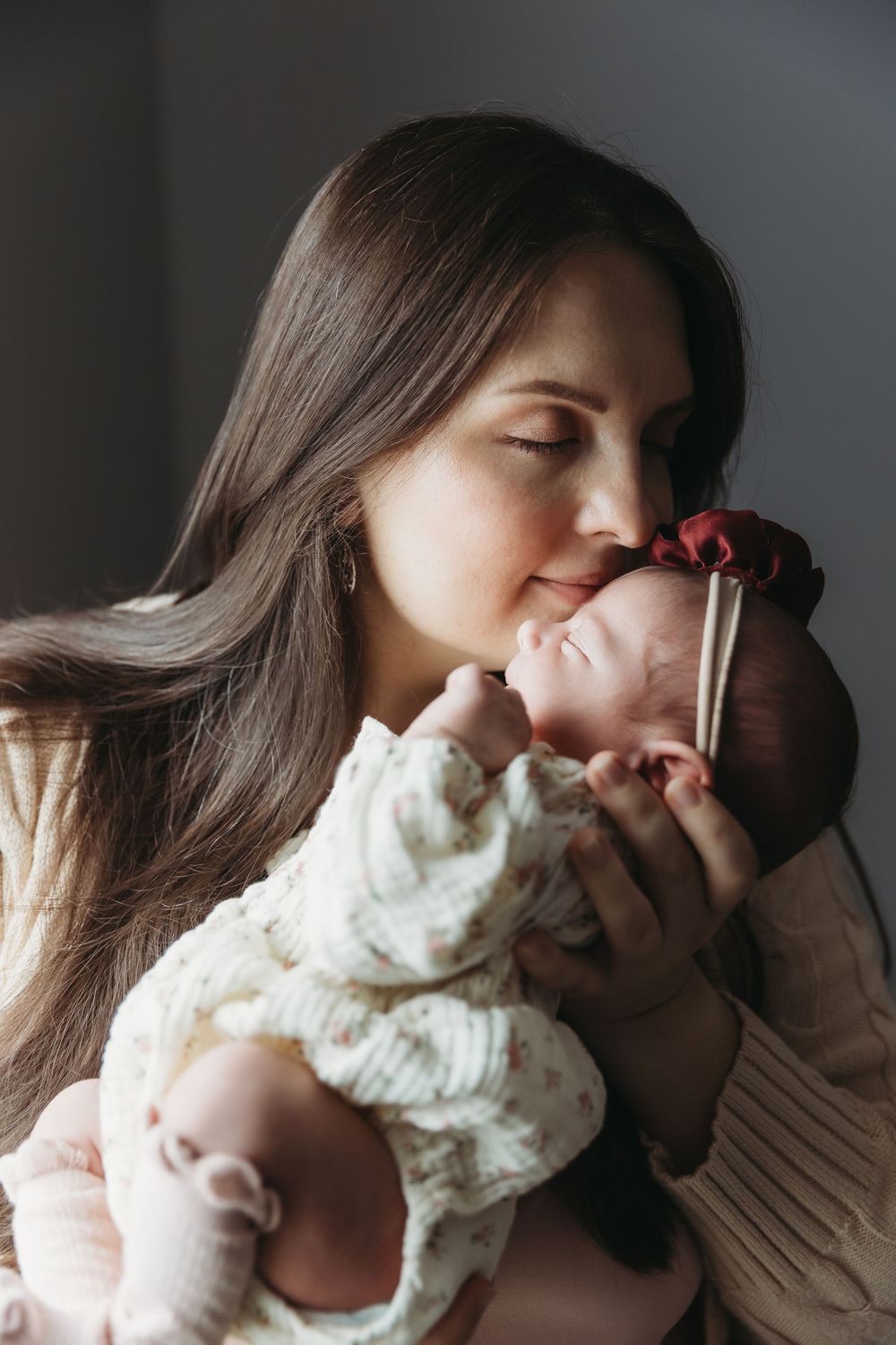 A young mother with long dark hair tenderly kisses her newborn baby dressed in a floral outfit with a red bow headband.