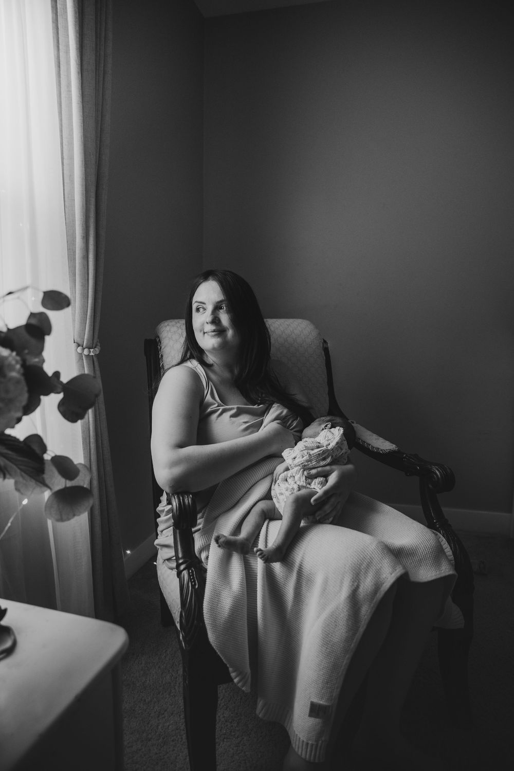 Black and white photo of a mother sitting in an armchair holding her newborn baby, bathed in soft window light.