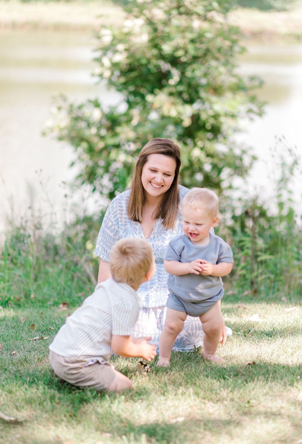 A smiling woman kneels on grass watching two young toddler boys play outdoors near a serene pond.