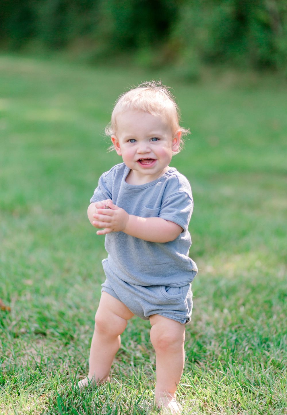 Happy toddler in gray romper standing on green grass, smiling outdoors in natural light.