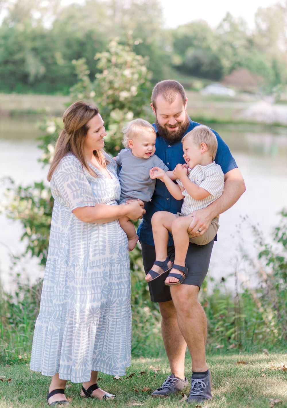 Family of four posing outdoors near a lake, parents holding two toddler boys, smiling together on a sunny day.