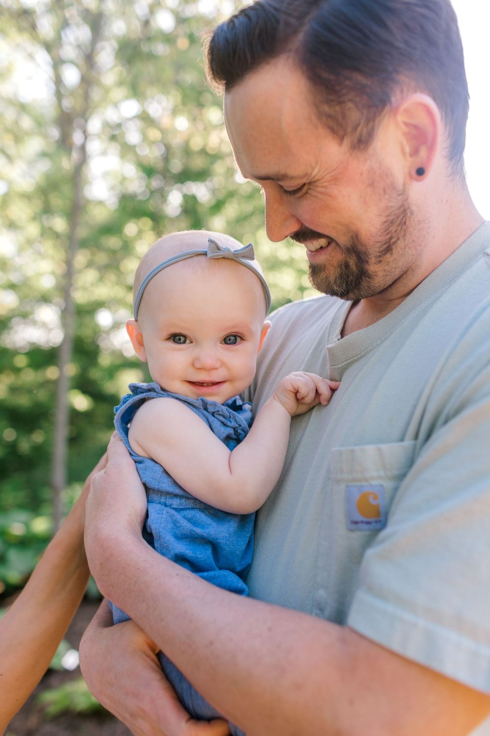 A bearded father smiles down at his happy baby girl in a blue romper and bow headband outdoors.