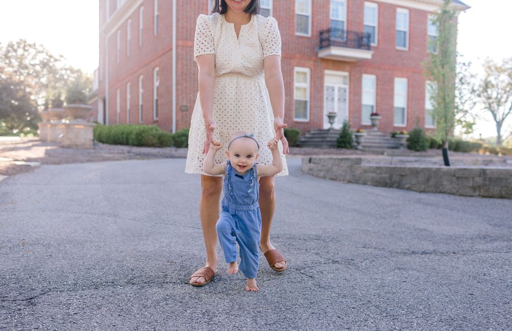 Mother helping baby take first steps on a brick building pathway, baby in denim overalls, mom in white dress.