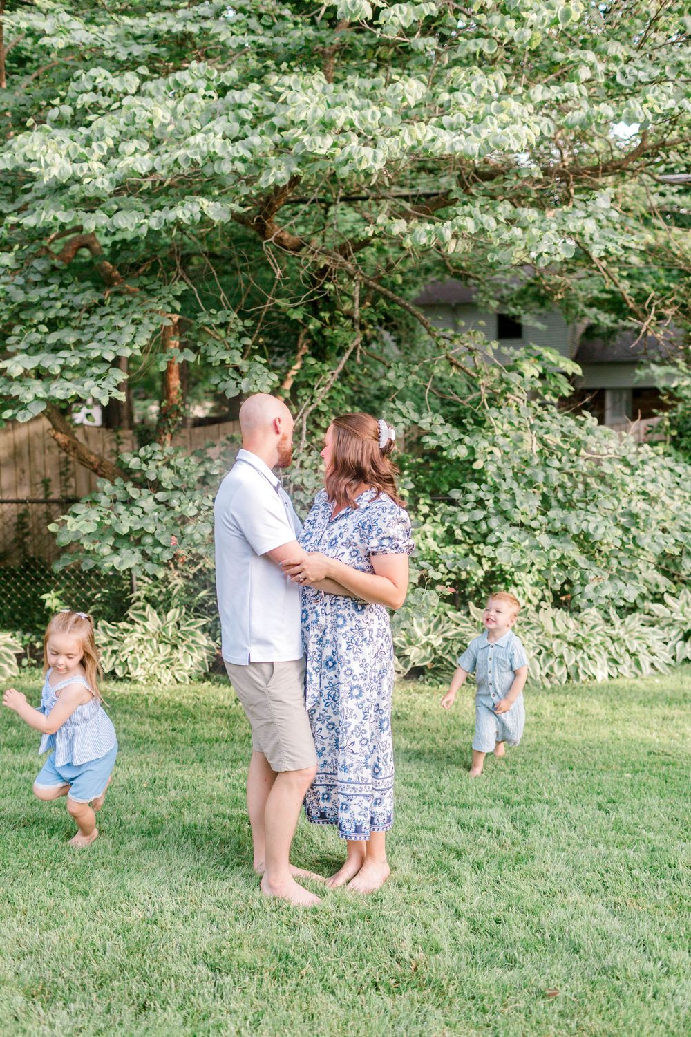 Family of four in a lush backyard, parents embracing while two young children play around them on green grass.