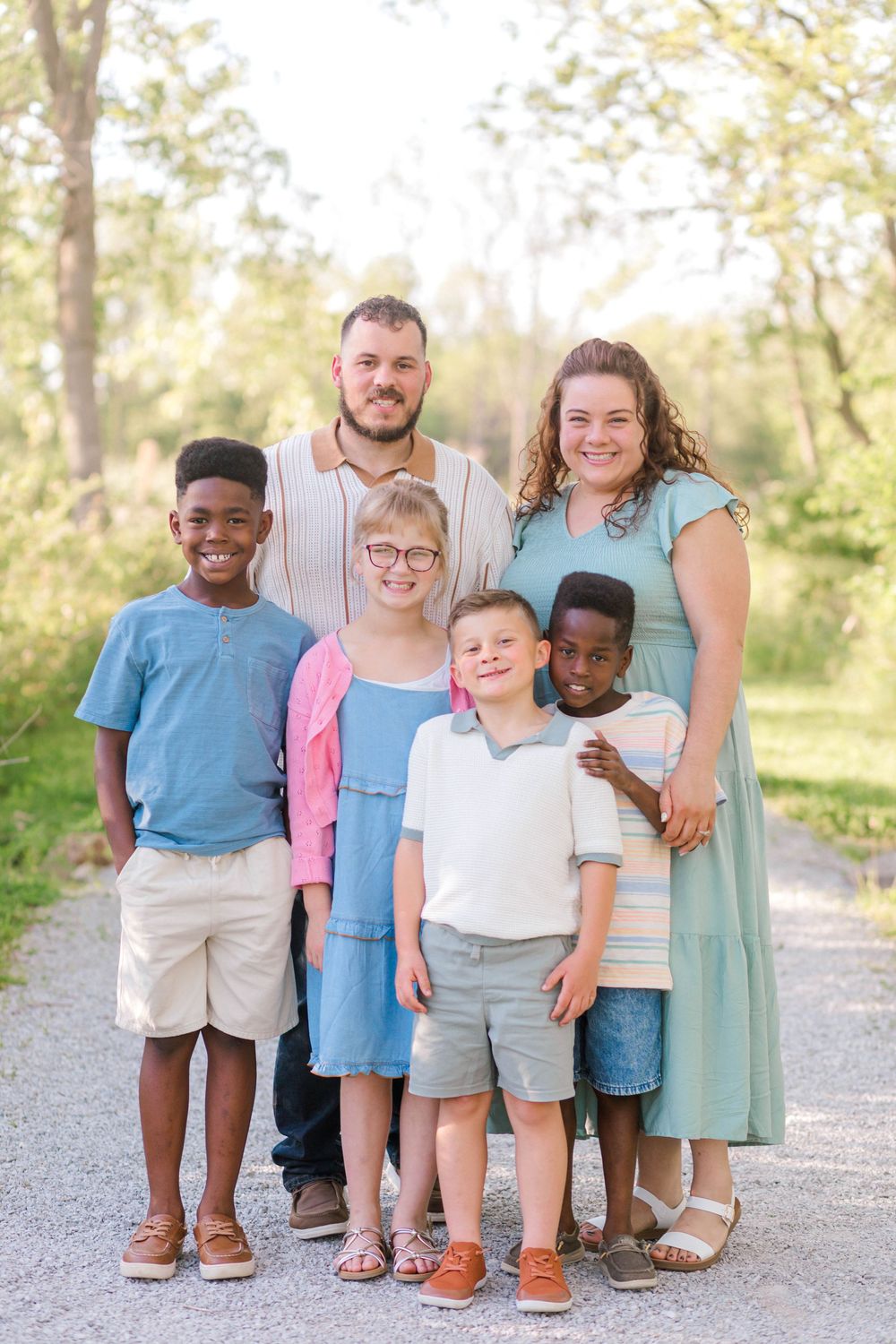 A joyful family of six poses outdoors on a sunny path, including two adults and four children of diverse backgrounds.