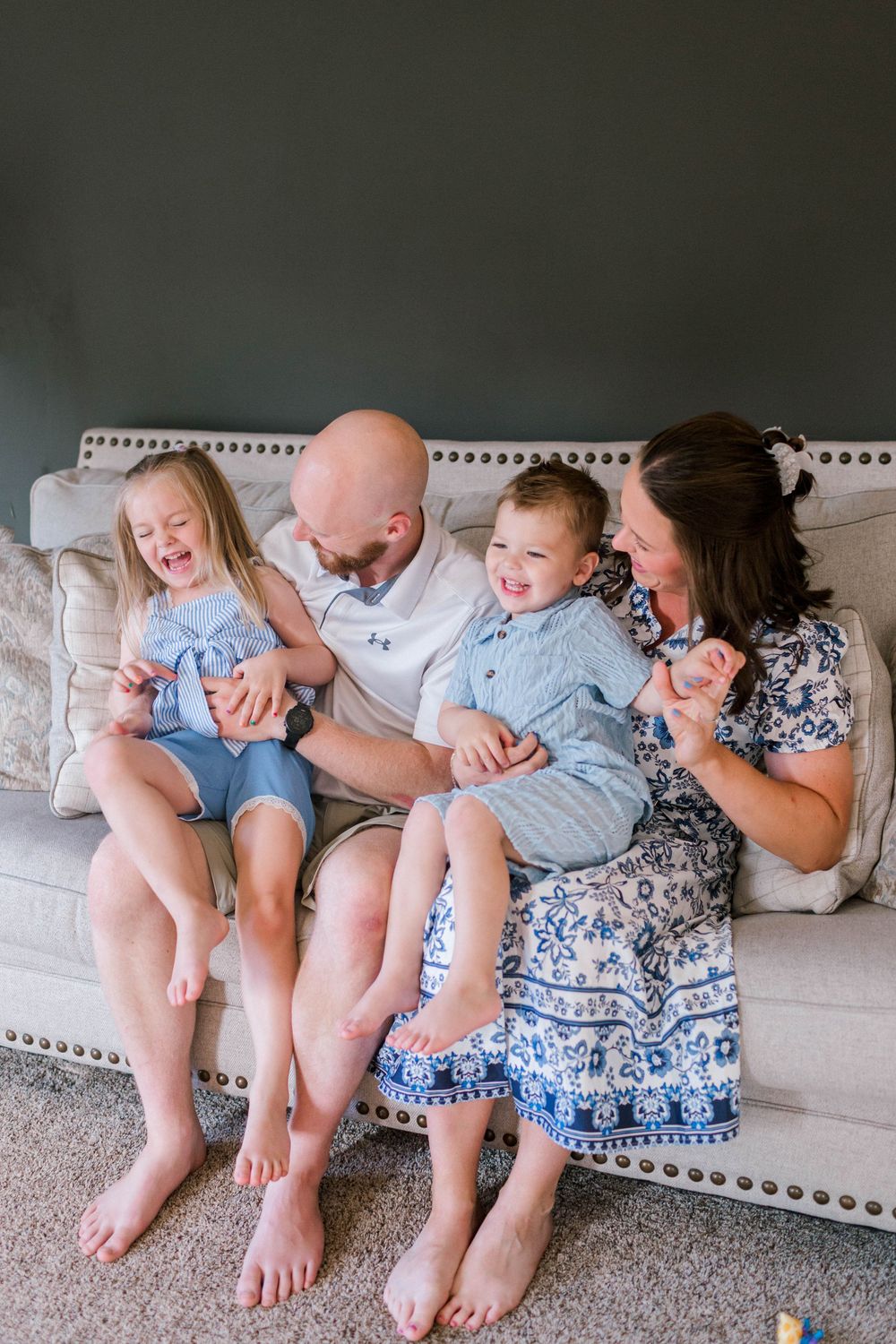 Happy family of four laughing together on a cream tufted sofa, parents tickling two young children in blue outfits.