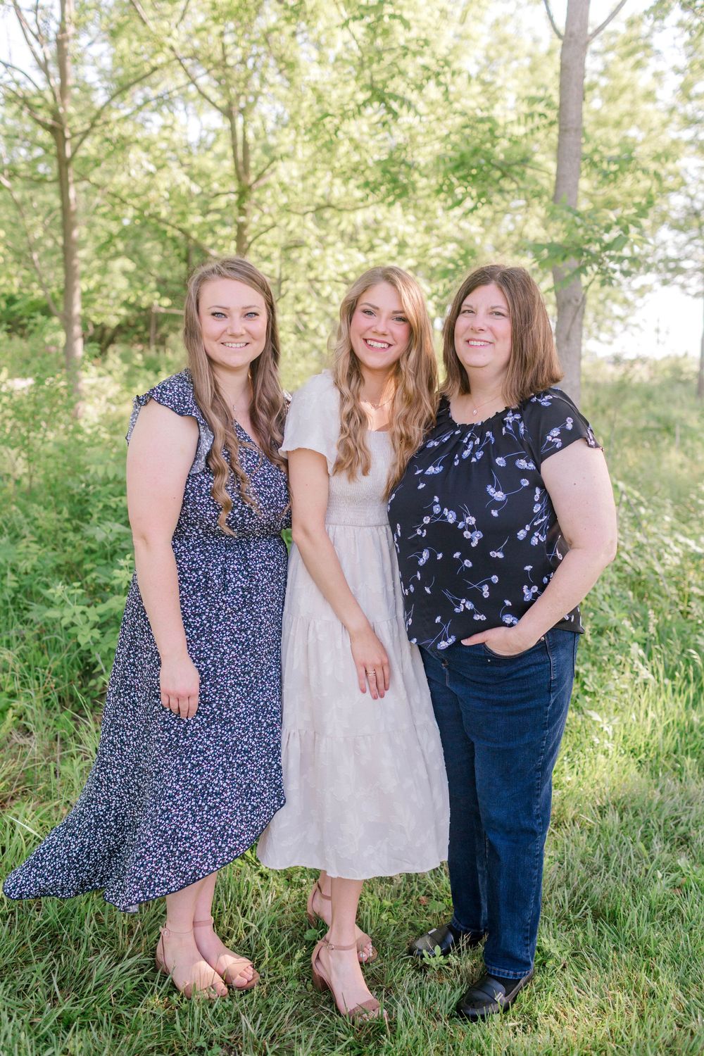 Three smiling women pose together outdoors in a lush green park setting, wearing summer dresses and casual attire.
