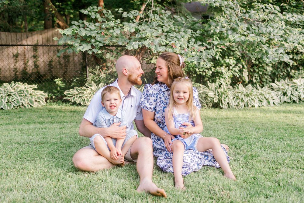 Happy family of four sitting on green grass in backyard, parents smiling at each other with two young children.