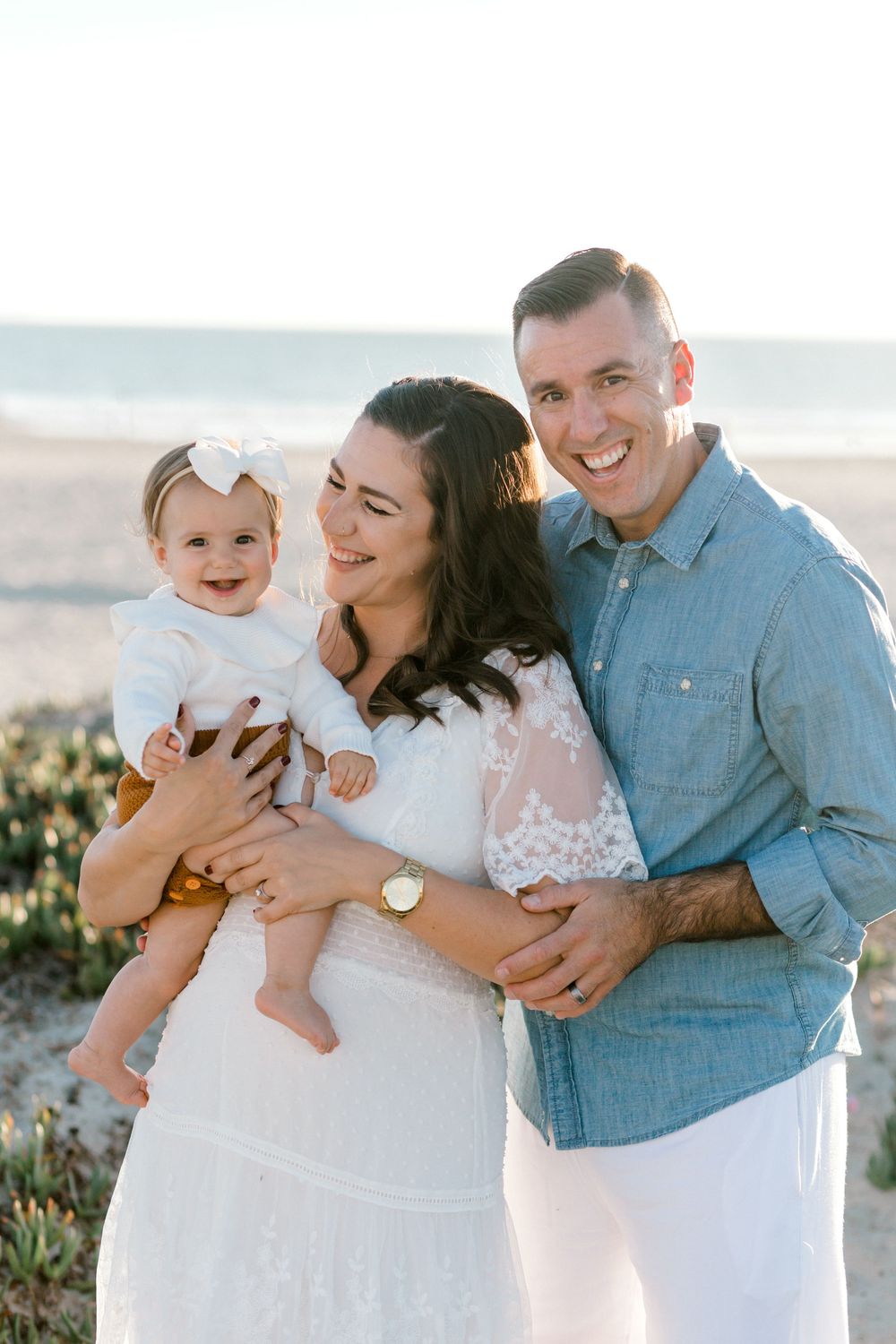 Happy family of three posing on beach, parents holding smiling baby girl in white bow, golden hour light.