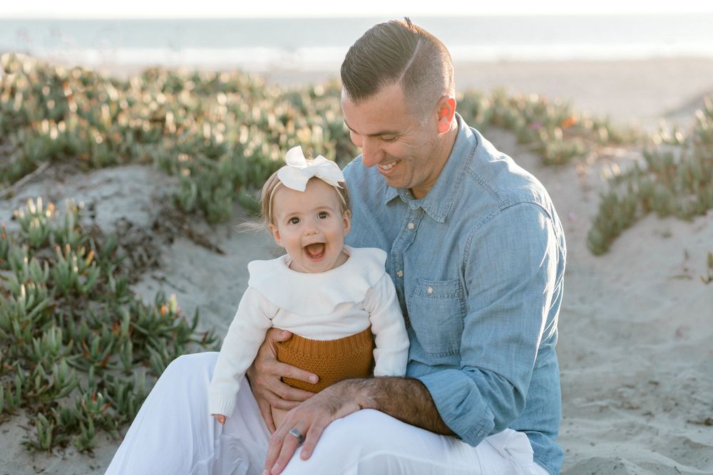 A smiling father holds his baby girl on a sunny beach, both dressed in white and denim, sitting on sand dunes.