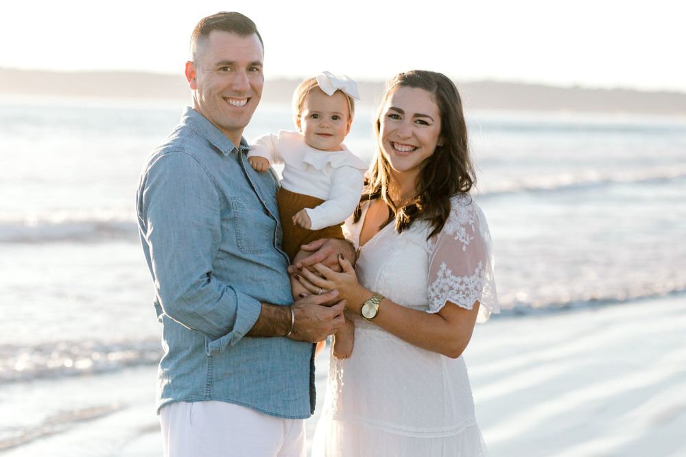 A smiling family of three poses on a sunny beach, with parents holding their baby girl in a white outfit.