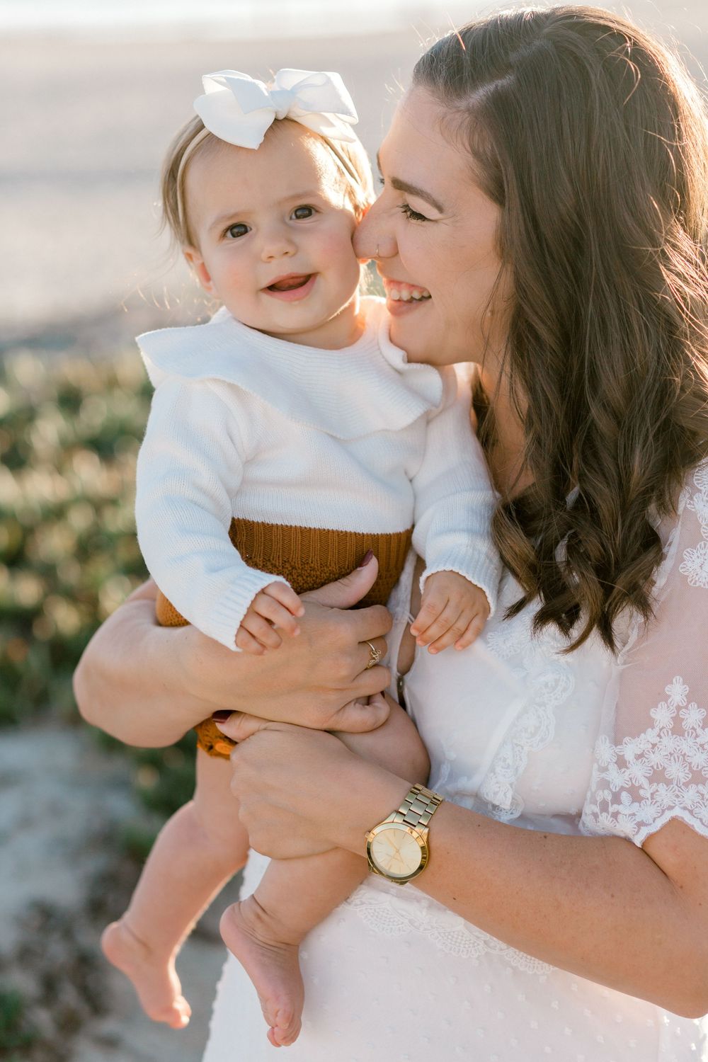 A smiling mother in white lace dress holds her happy baby girl wearing a white top and bow on a sunny beach.