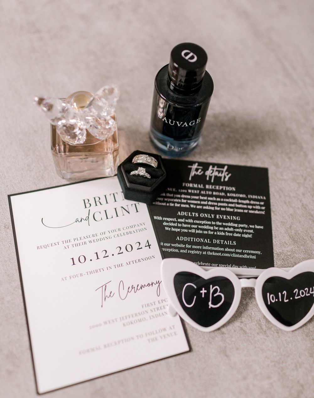 Wedding flat lay featuring invitation suite, rings, Dior cologne, perfume, and heart sunglasses.