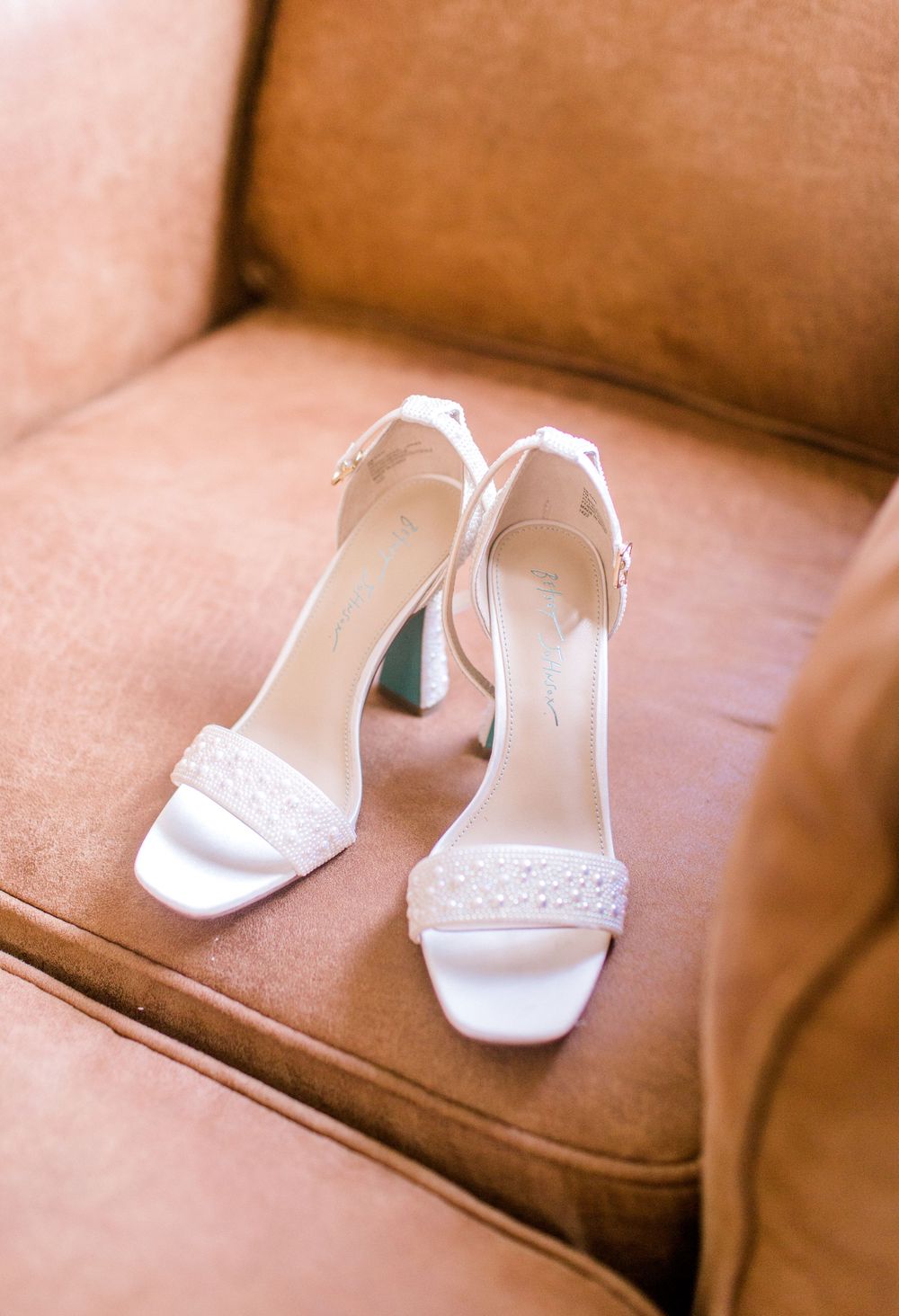 Elegant white bridal block heels with pearl embellished straps resting on brown leather chair.