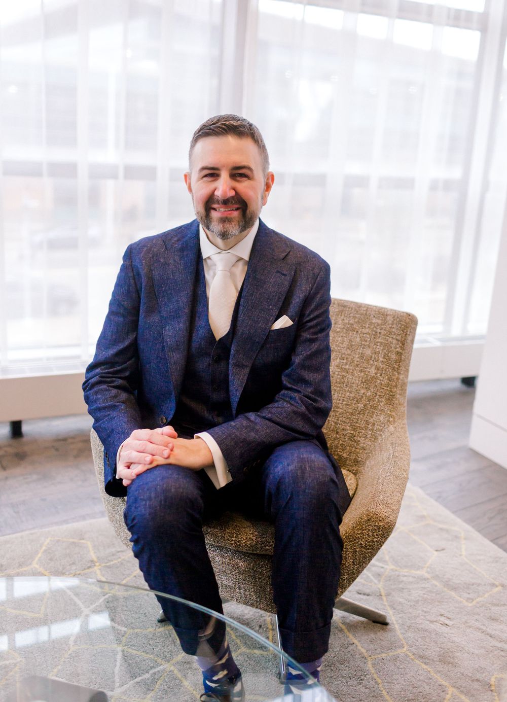 Smiling groom in navy blue three-piece suit seated in a modern chair by large windows.