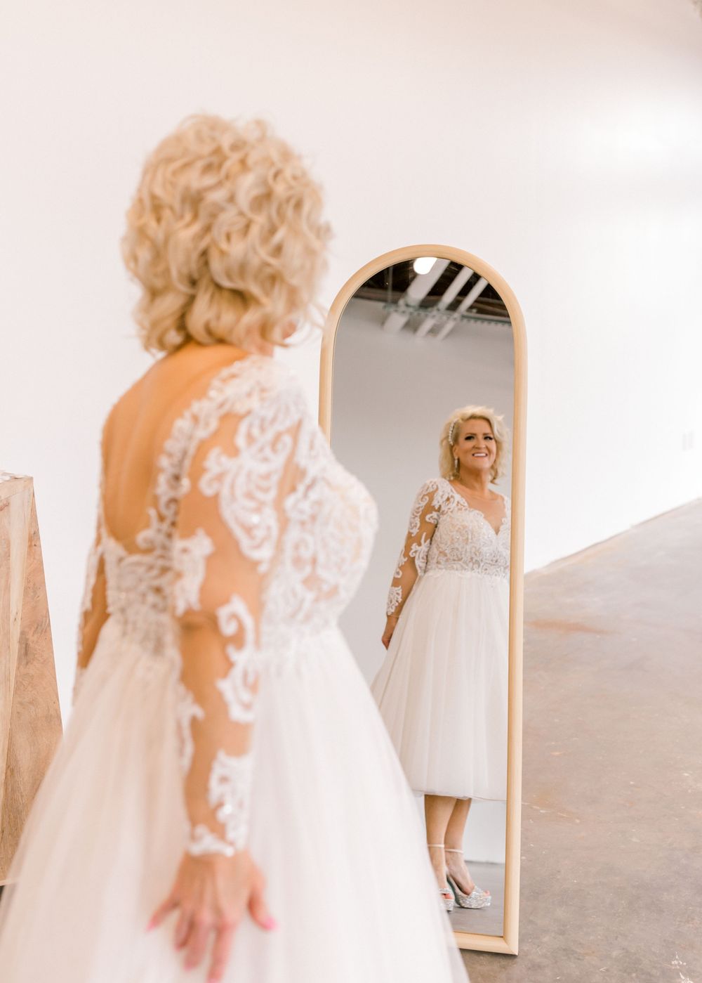 Blonde bride in lace long-sleeve wedding gown admiring her reflection in an arched mirror.