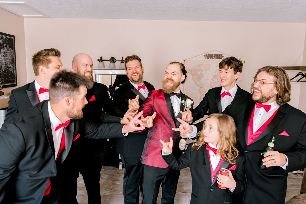 Groom in red velvet jacket celebrates with groomsmen in black tuxedos and red accessories.