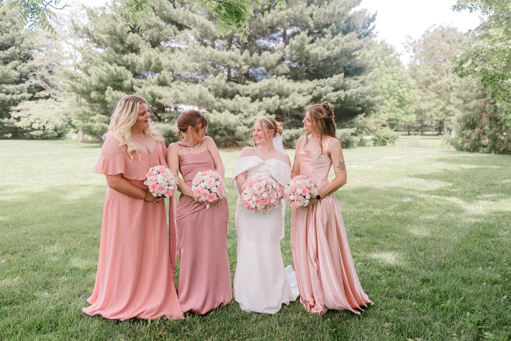 Bride in white gown with three bridesmaids in pink dresses holding bouquets on green lawn at outdoor wedding.