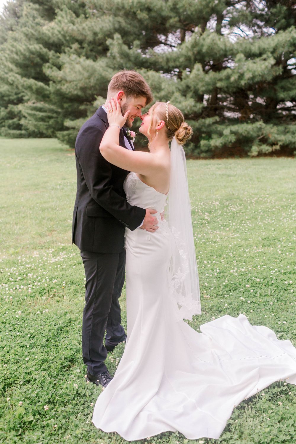 Bride and groom share a romantic forehead touch in a white gown and black suit on a lush green lawn.