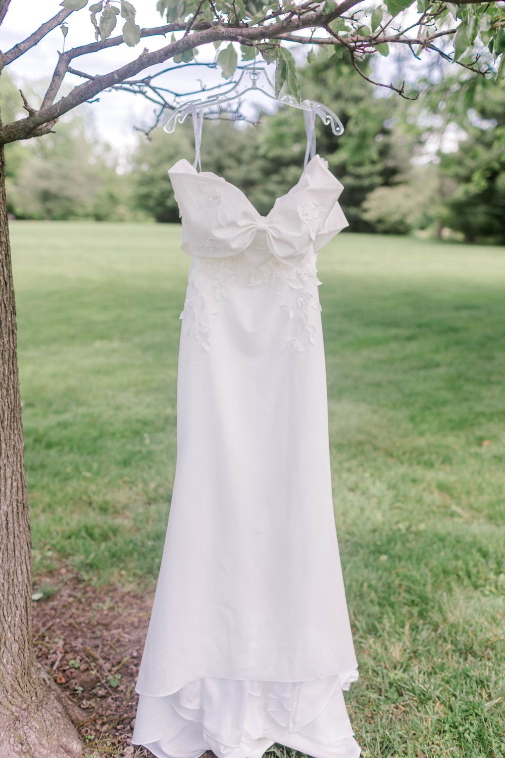 White wedding dress with floral appliques hanging from a tree branch outdoors in a lush green garden.