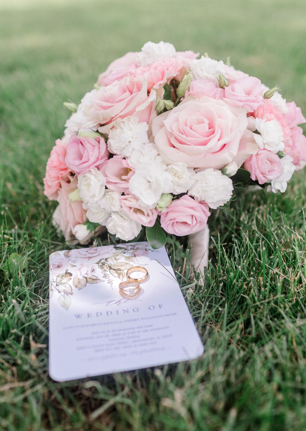 Pink and white rose wedding bouquet with gold rings on a wedding program card, resting on green grass.