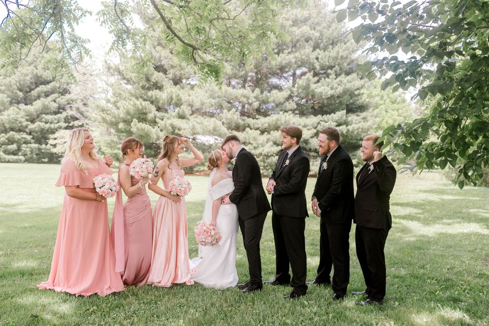 Wedding party poses outdoors, bridesmaids in pink gowns and groomsmen in black suits flanking the kissing bride and groom.