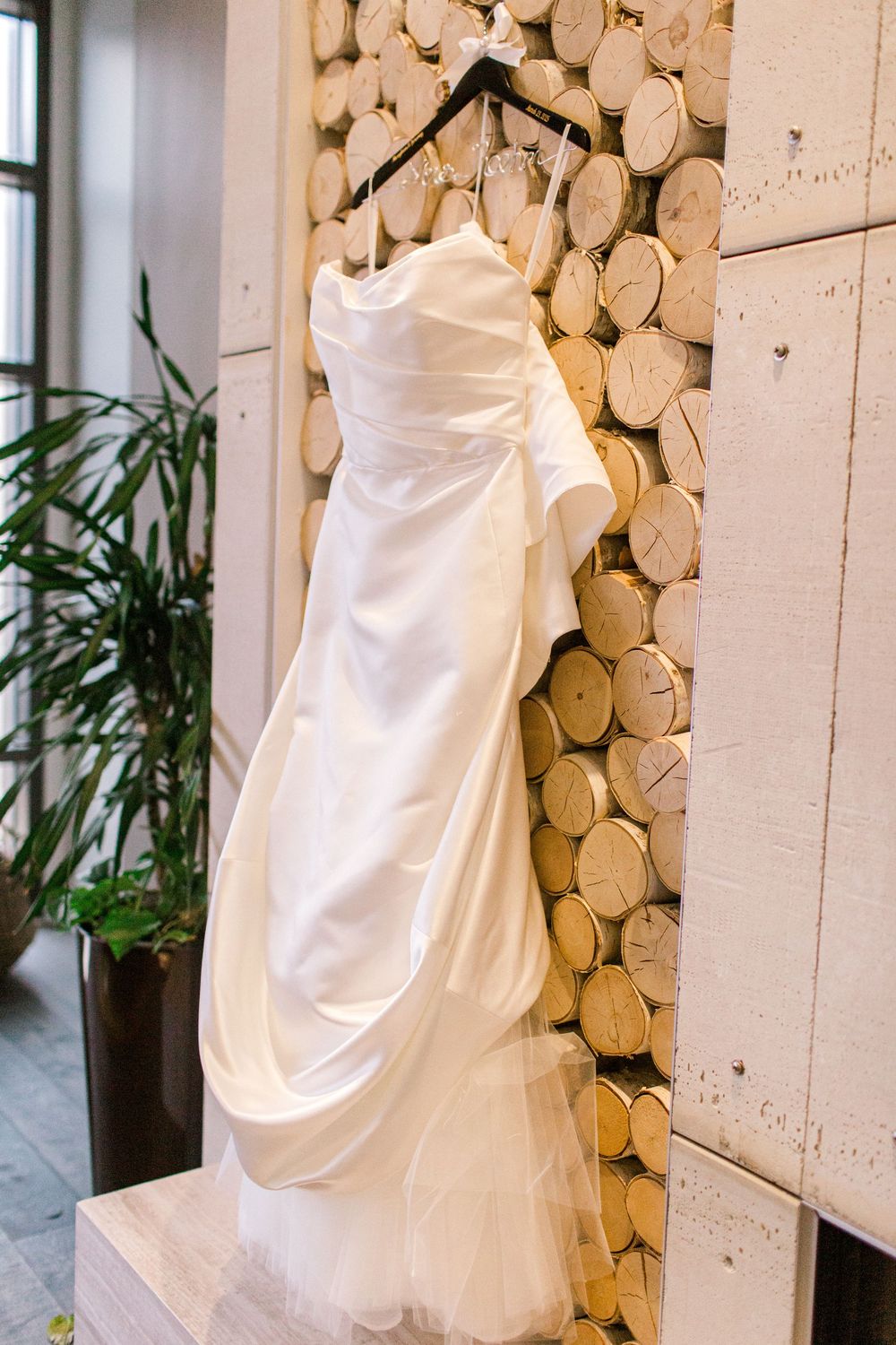 Sleek white strapless wedding gown hanging on hanger against rustic wood log wall display.