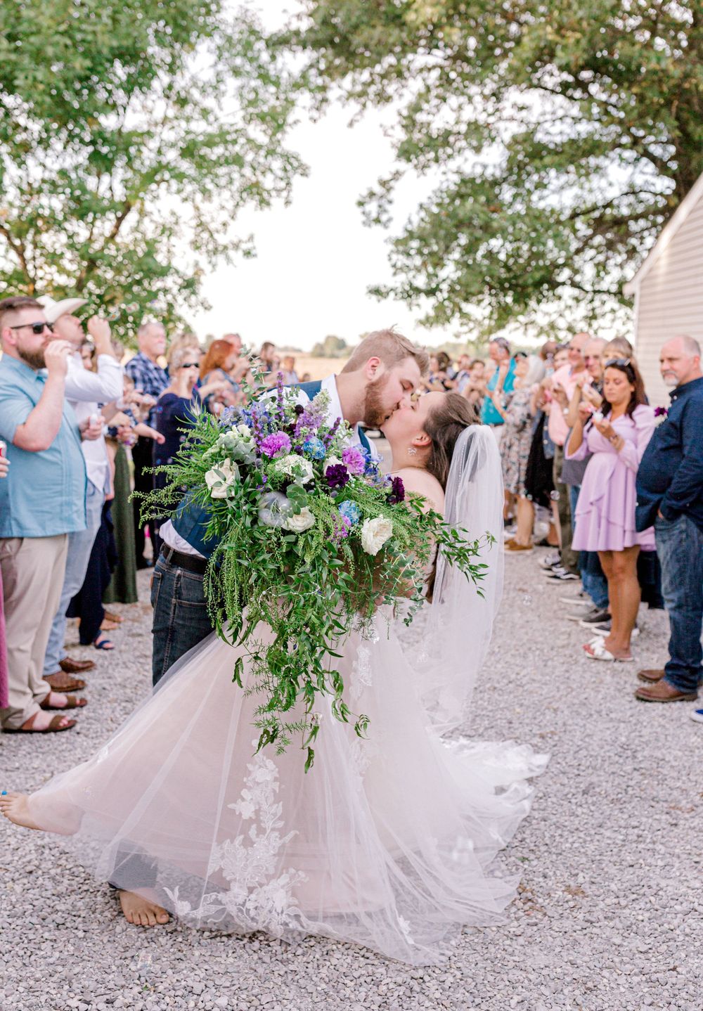 Bride and groom sharing a romantic dip kiss surrounded by cheering guests at outdoor wedding.