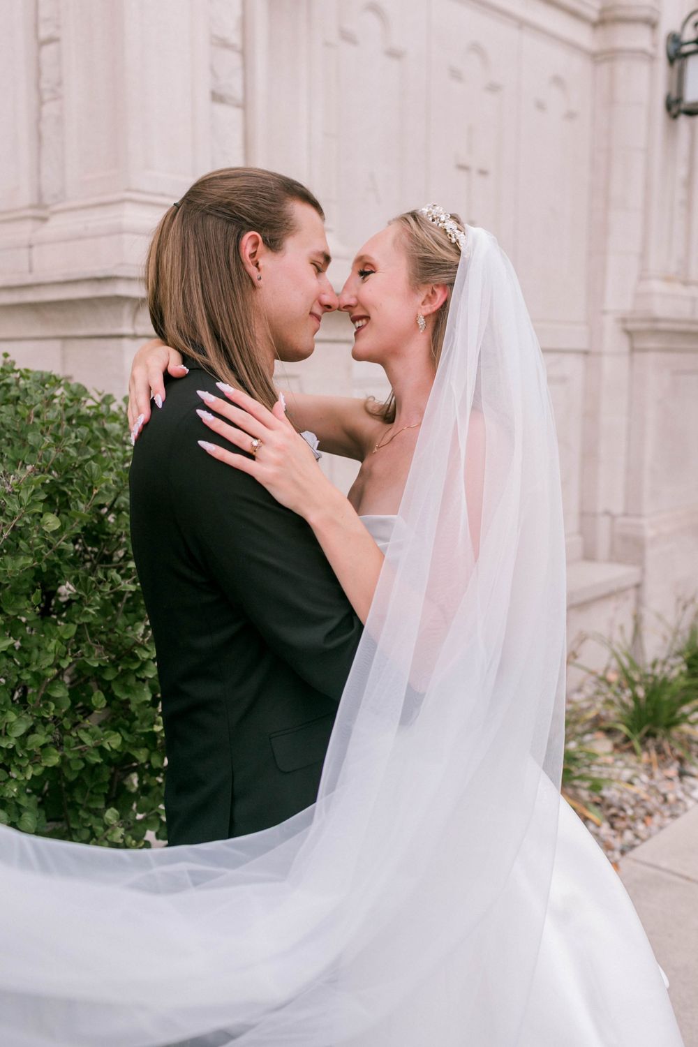 Bride and groom share a romantic nose-to-nose moment outdoors, her flowing veil billowing as they embrace near a stone building.