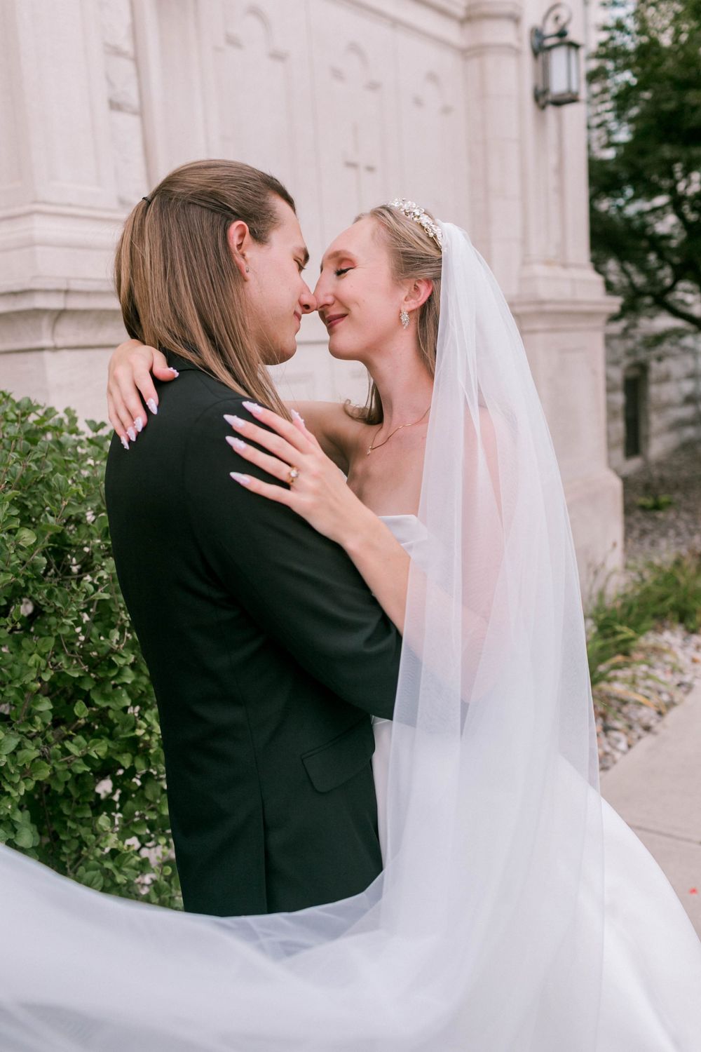 A bride in a white gown and veil shares a romantic moment with her groom in a black suit outdoors.