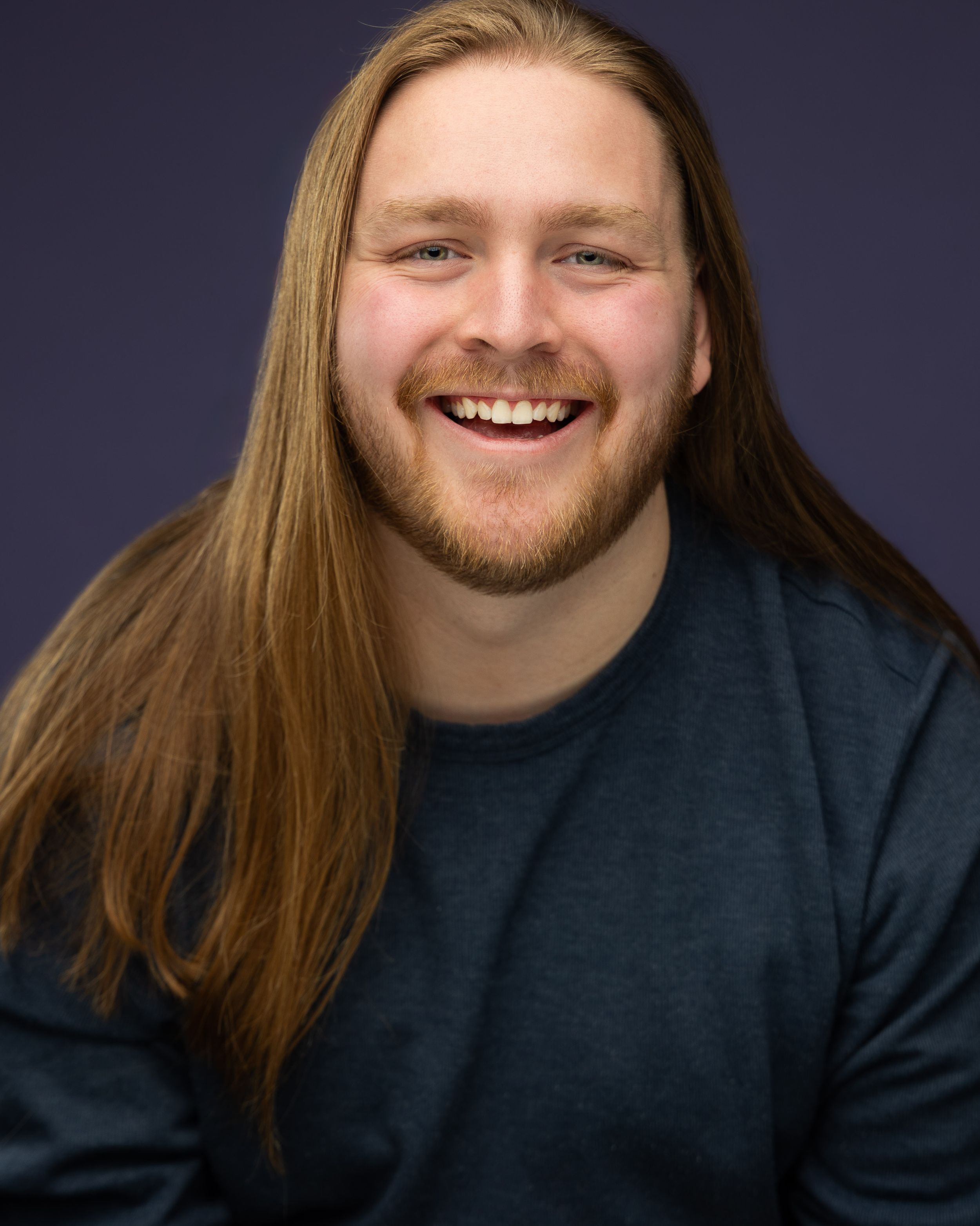 Smiling young man with long auburn hair and beard wearing a dark teal shirt against a purple background.