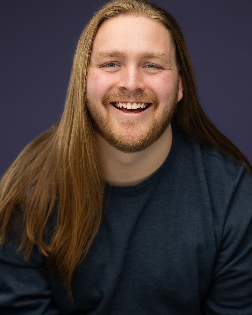 Smiling young man with long auburn hair and beard wearing a dark teal shirt against a purple background.