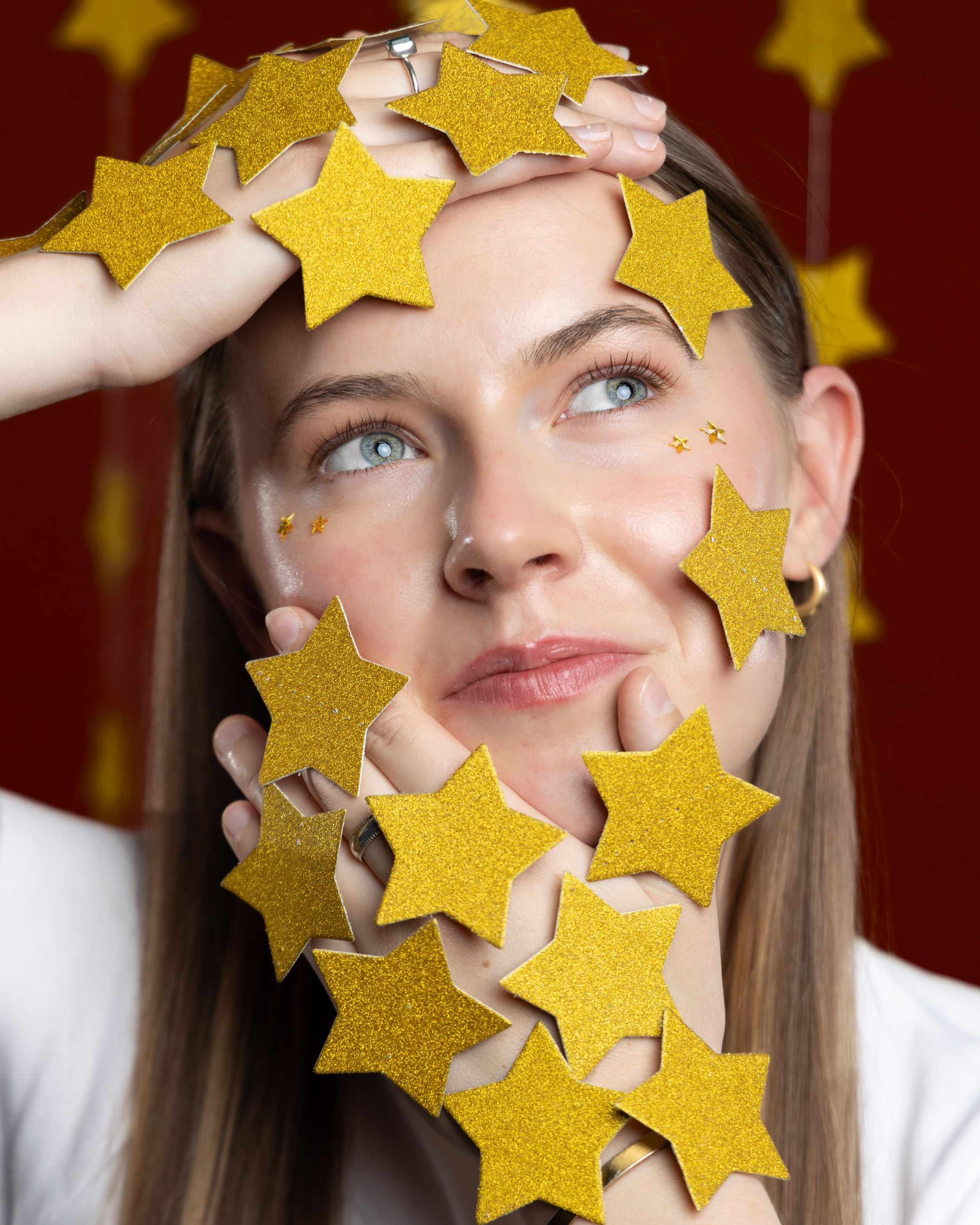 Young woman with blue eyes surrounded by glittery gold stars on a red background, looking up dreamily.