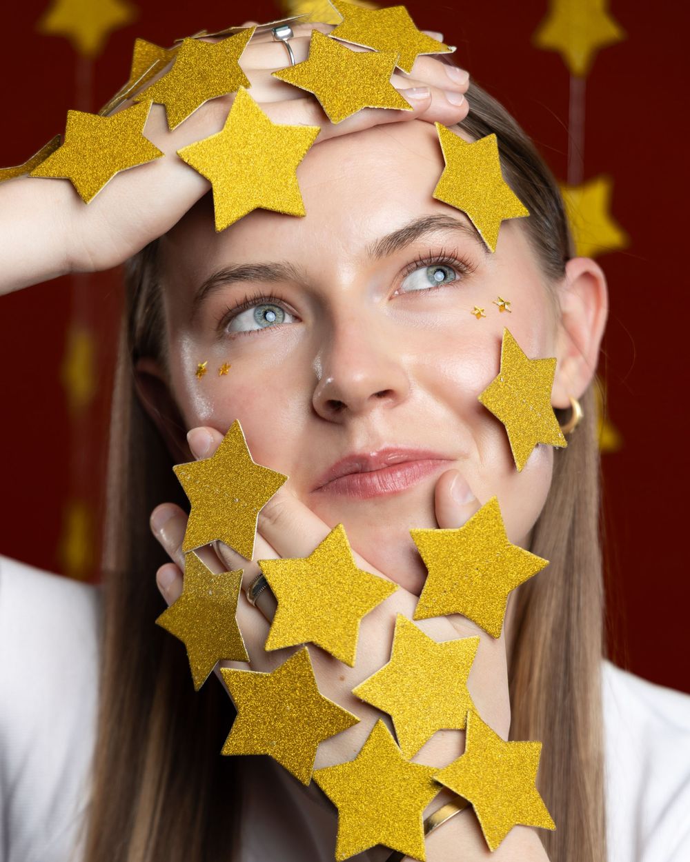 Young woman with blue eyes surrounded by glittery gold stars on a red background, looking up dreamily.