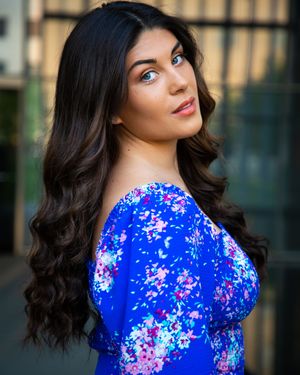 Headshot showing a woman in a vibrant blue floral dress against urban background.
