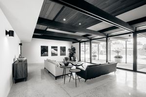 Modern open concept living space with black ceiling beams, floor-to-ceiling windows, and minimalist white decor.