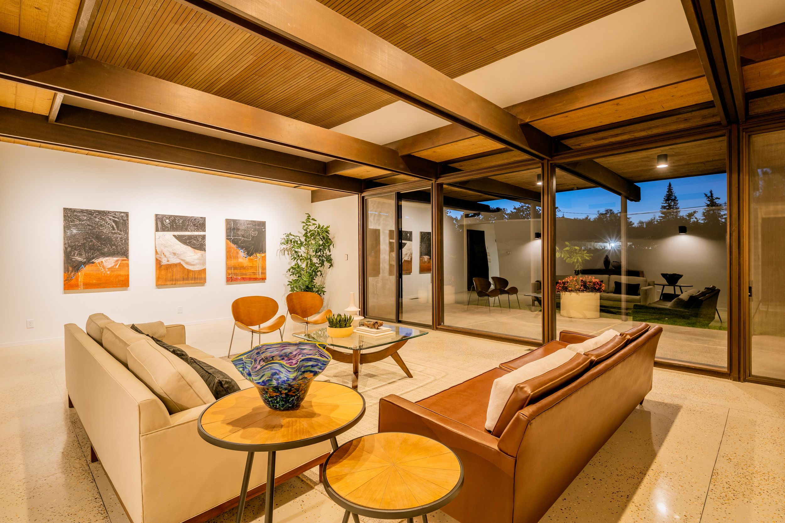 Modern living room with exposed wood beams, glass walls, and mid-century modern furniture in warm earth tones.