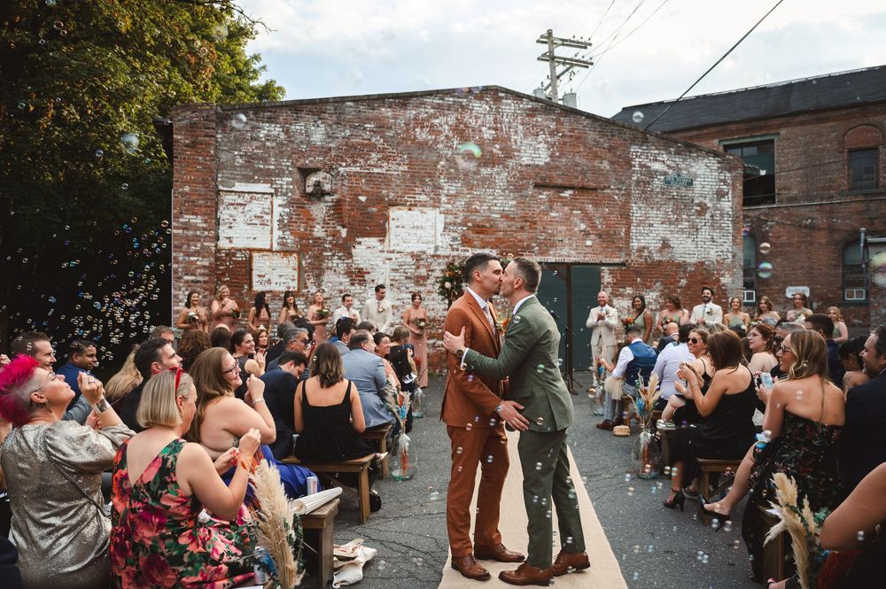Grooms kiss surrounded by applauding guests in front of industrial wall and arch with flowers, at Garner Arts Center in the Hudson Valley.