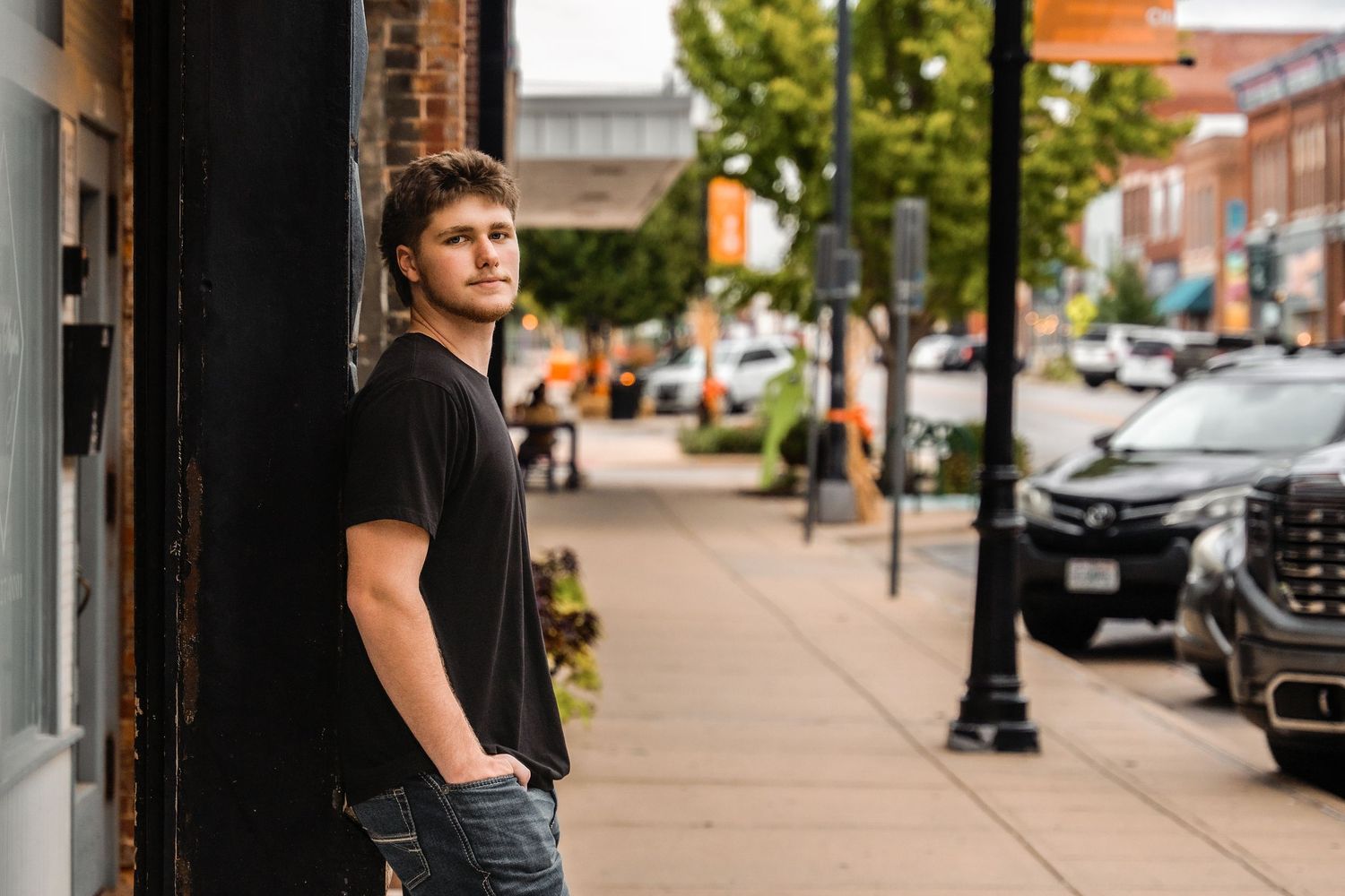 A person in a black t-shirt stands against a brick wall in an urban setting with storefronts and parked cars visible.
