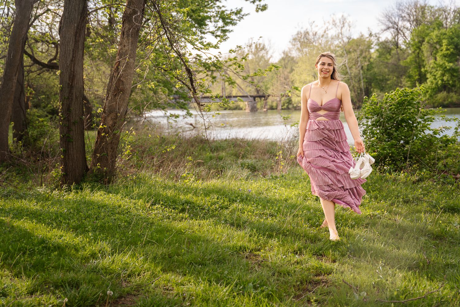 A woman in a pink dress walks on green grass near a river with trees and a bridge in the background.
