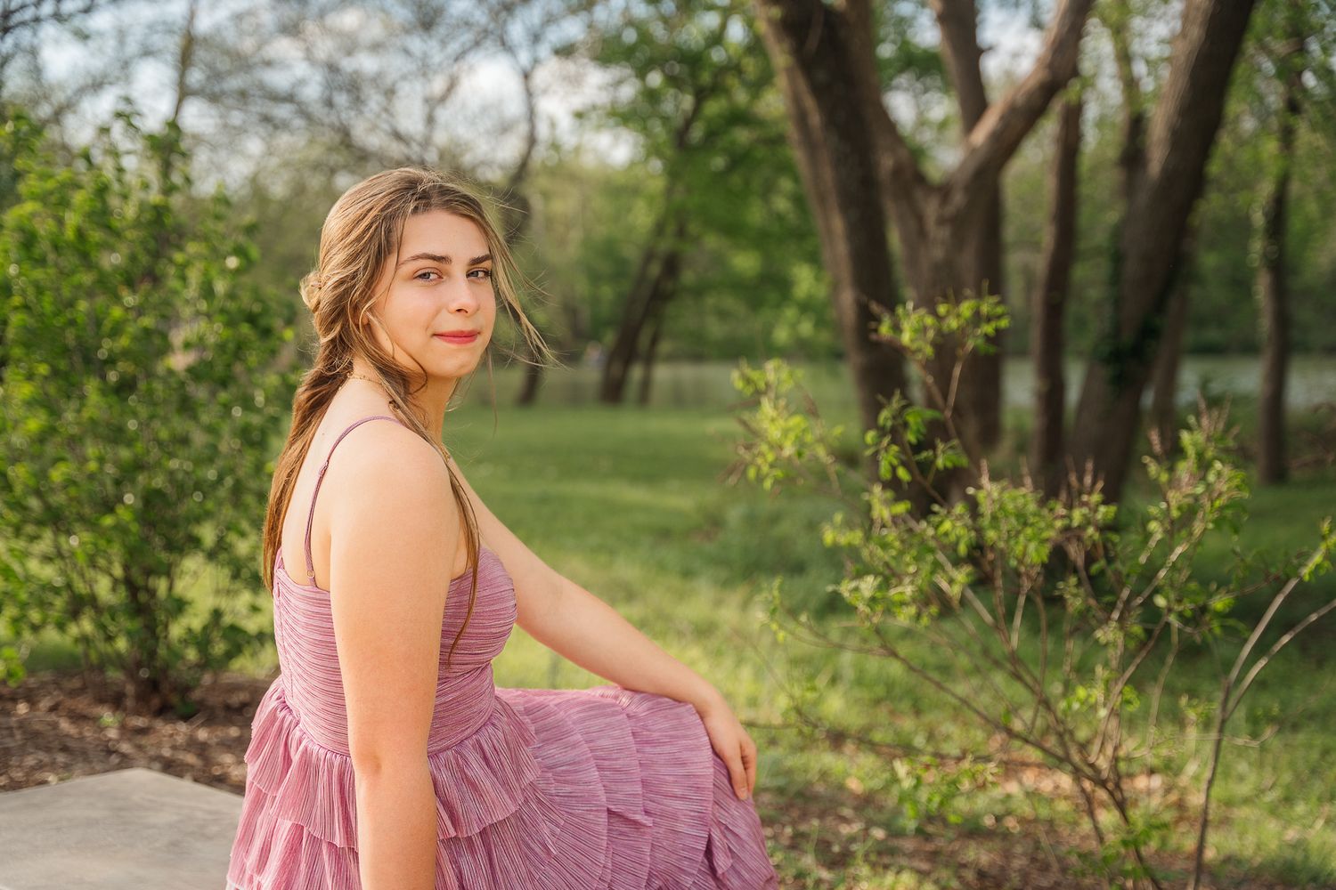 Young woman in pink dress sitting outdoors in lush green park, smiling over shoulder in natural light.