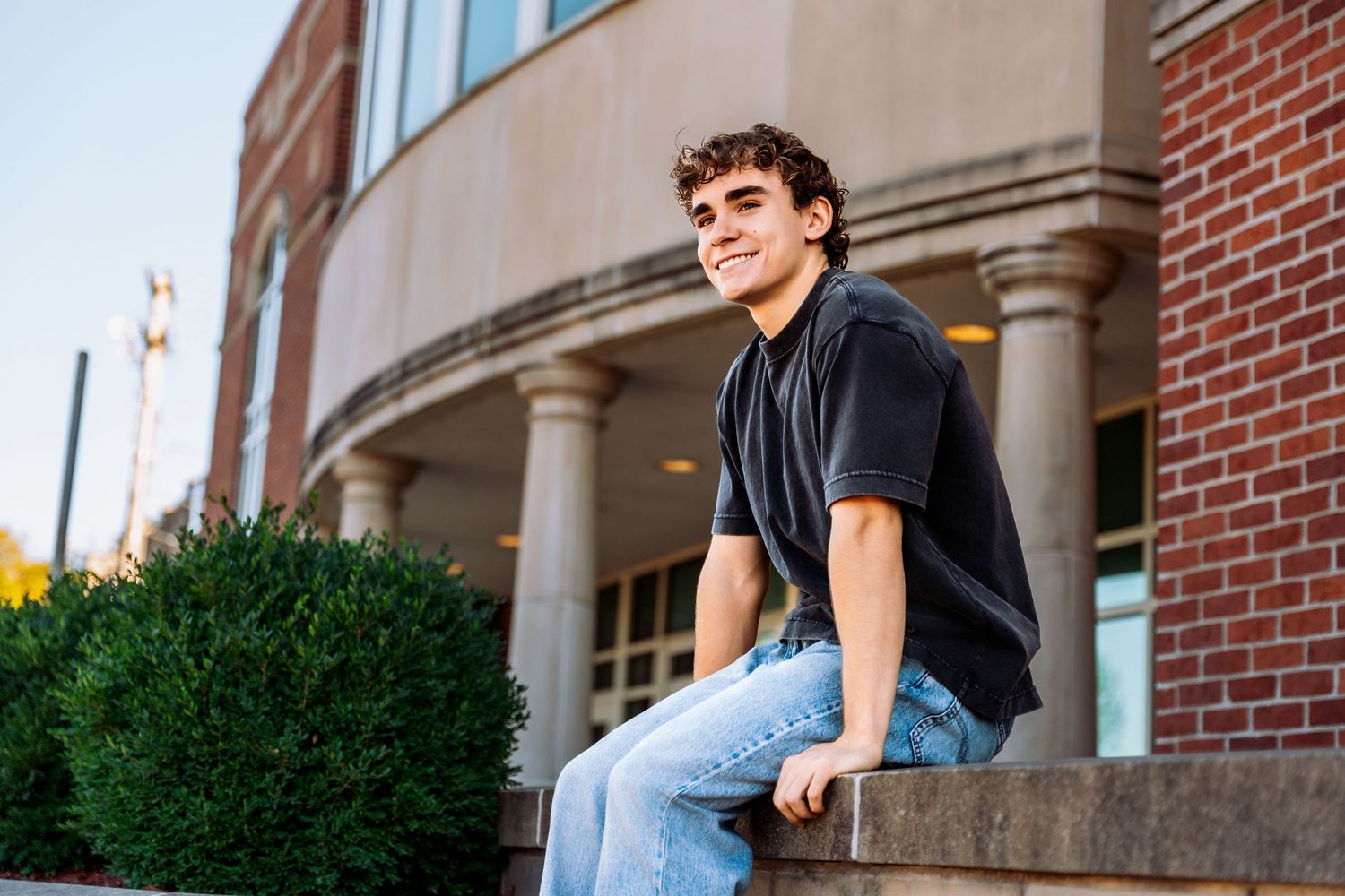 Student in casual attire sits thoughtfully on stone steps of a brick building on a college campus.
