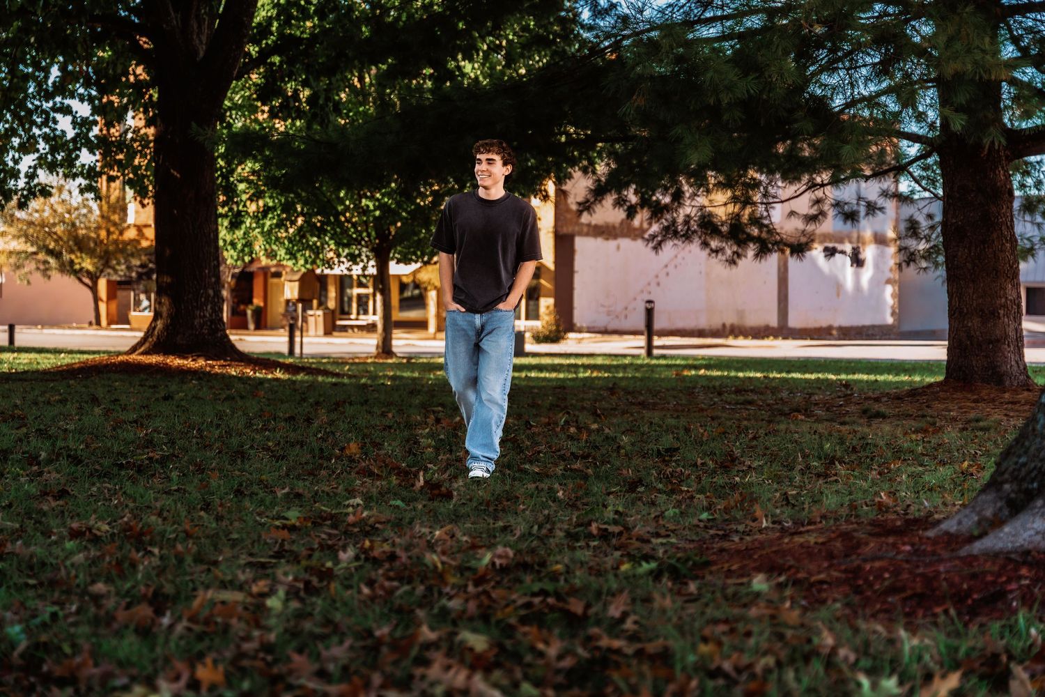 A person walks on grass beneath tall trees at sunset with a building visible in the background.