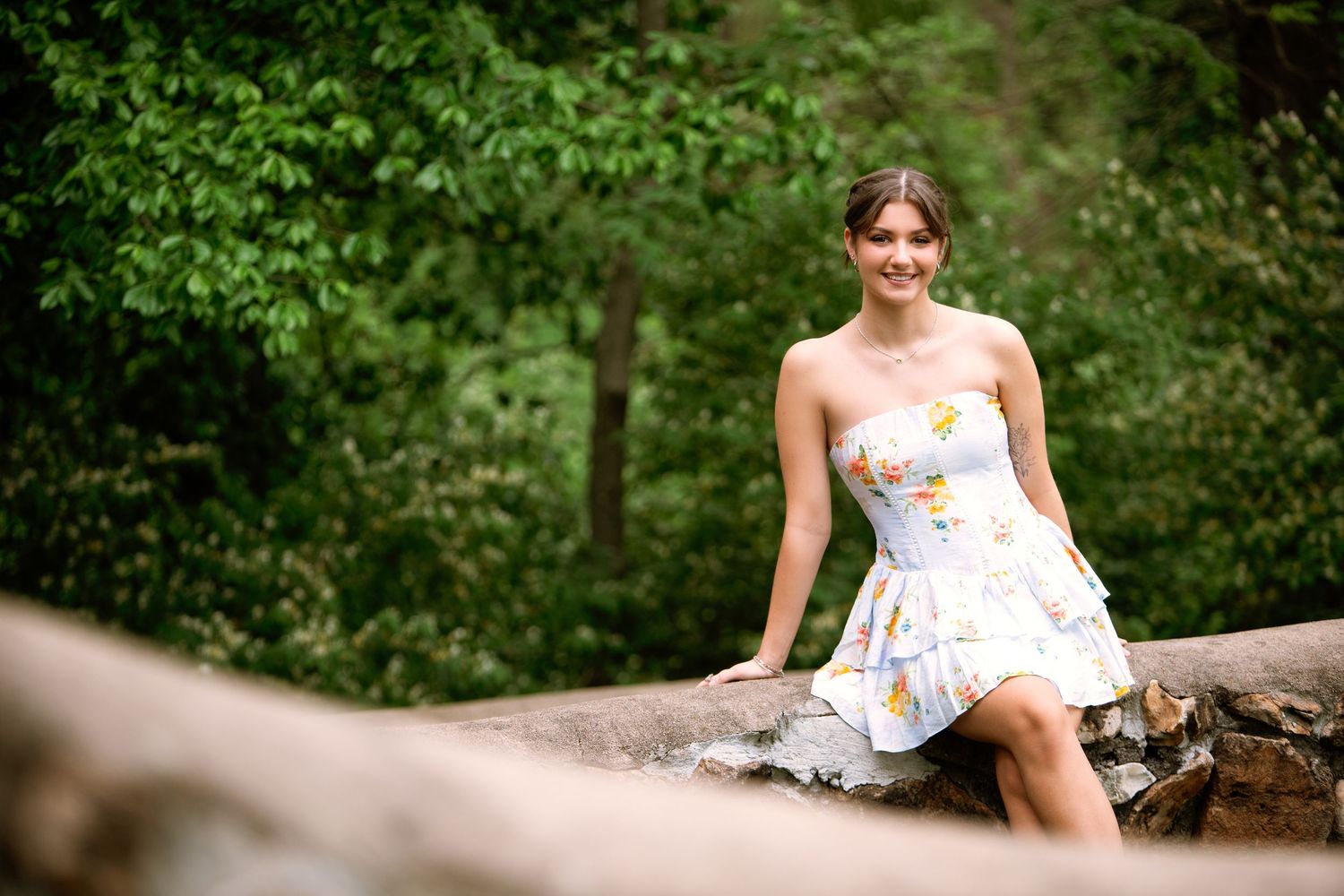 A person in a white floral dress sits on a fallen tree trunk in a lush green forest setting.