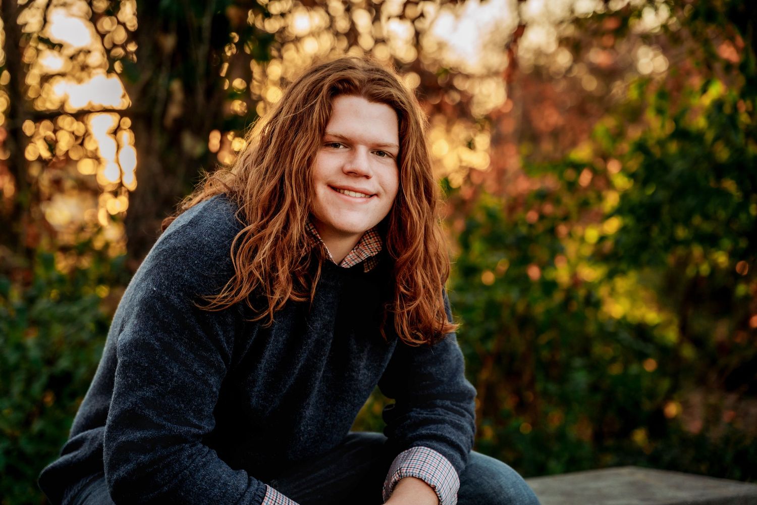A person with long brown hair sits outdoors during golden hour, surrounded by autumn foliage.