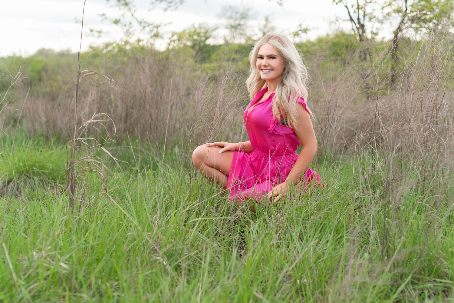 A flowing pink dress creates an ethereal effect against a natural backdrop of tall grass and trees.