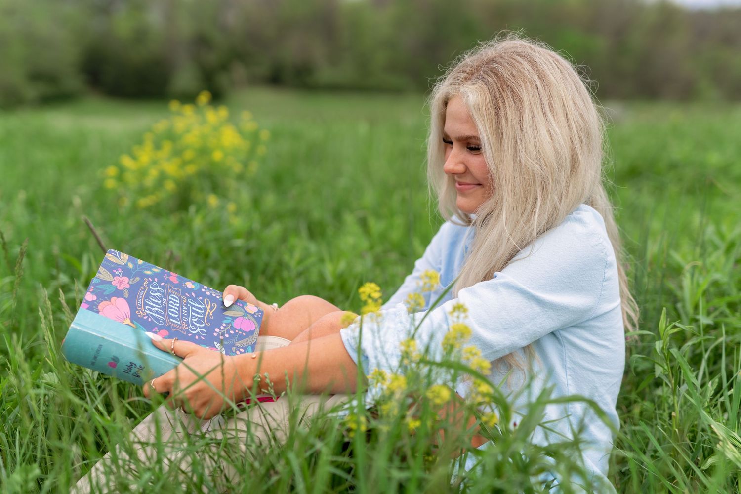 A person in a light blue shirt lies in a green meadow reading a book among spring wildflowers.